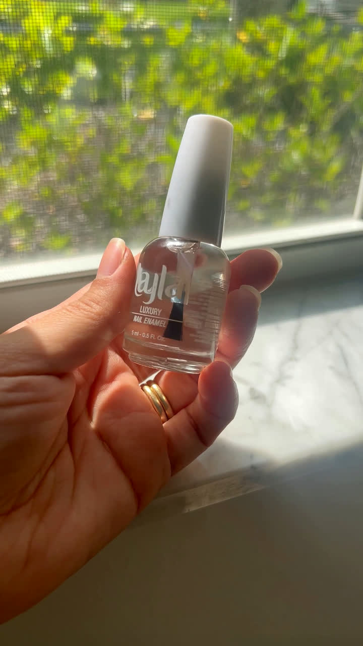 Hand holding a clear Layla nail enamel bottle by a window with greenery outside